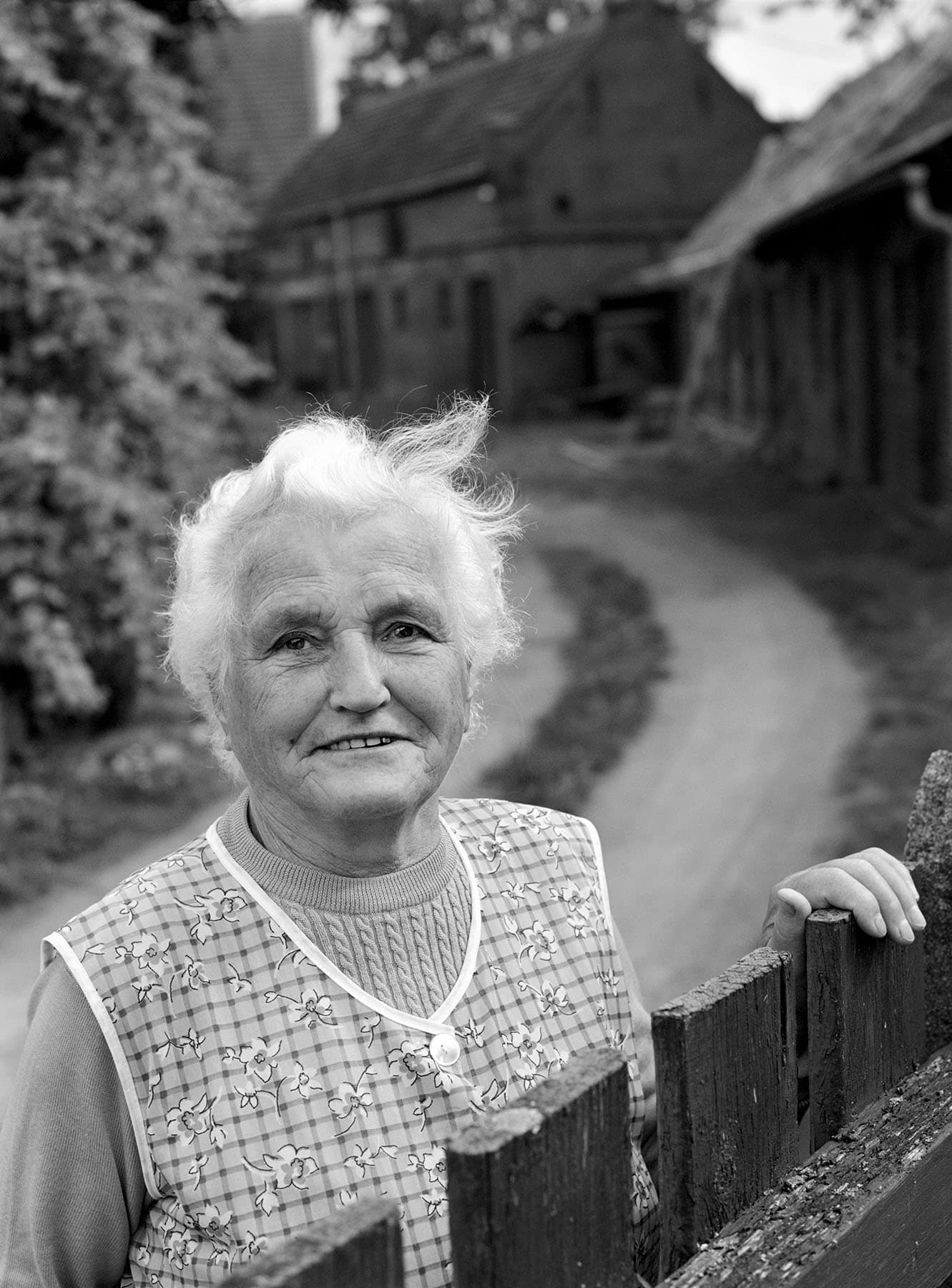 Portrait ältere Frau am Zaun und Fachwerkhäusern aus dem Fotoprojekt Ein Dorf in unserer Zeit – Menschen in ihrer dörflichen Umgebung von Uwe Nölk